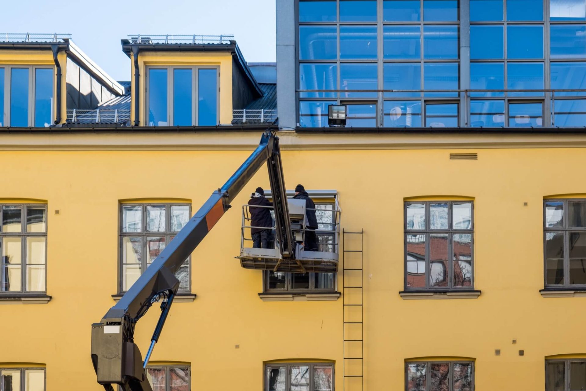 En ljusgul fastighet som genomgår en renovering, med två byggarbetare som arbetar på fasaden från en höjande plattform