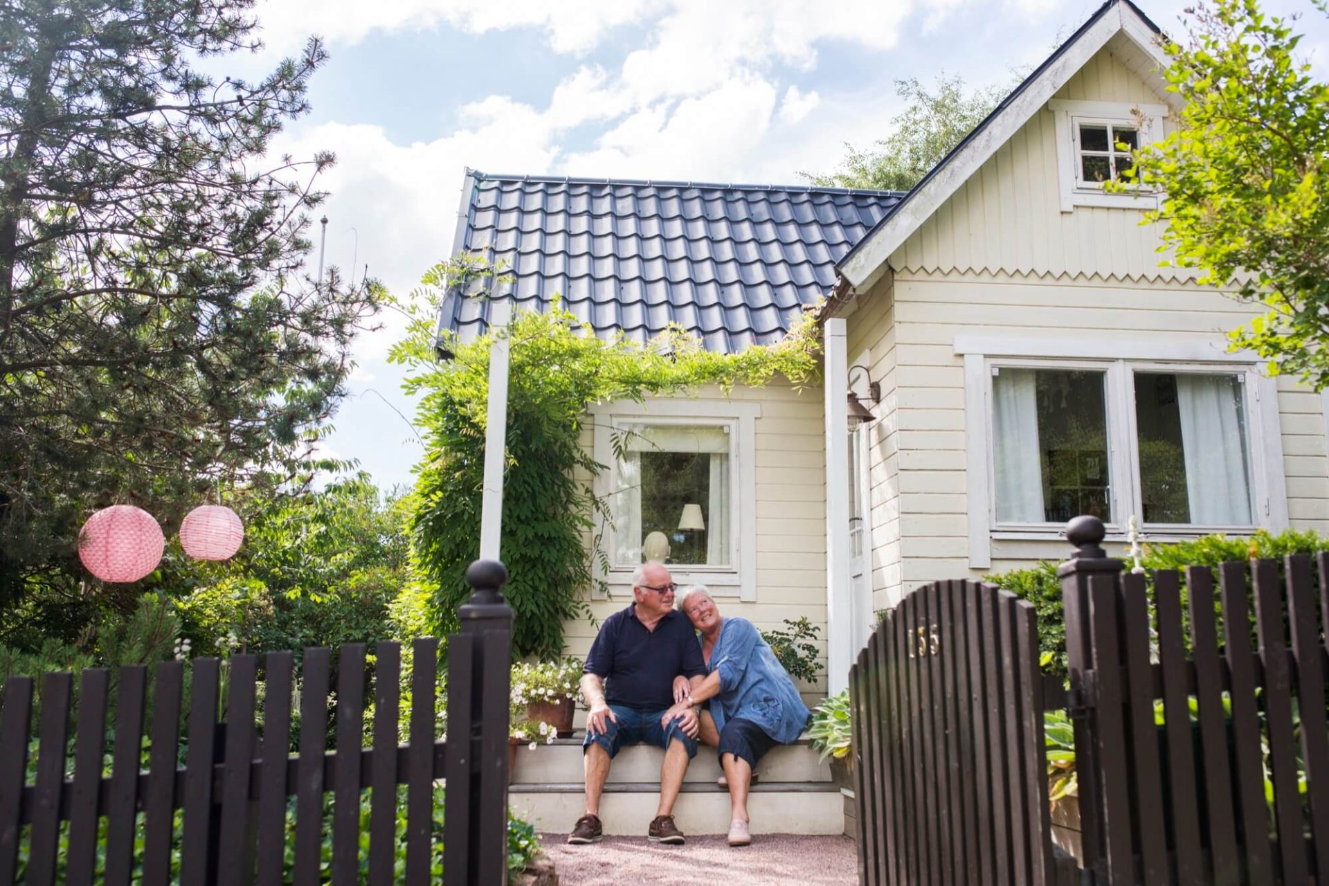 Ett äldre par finner sin fristad på verandan till deras ljusgula trähus med svart tak, omslutna av ett hav av grönska